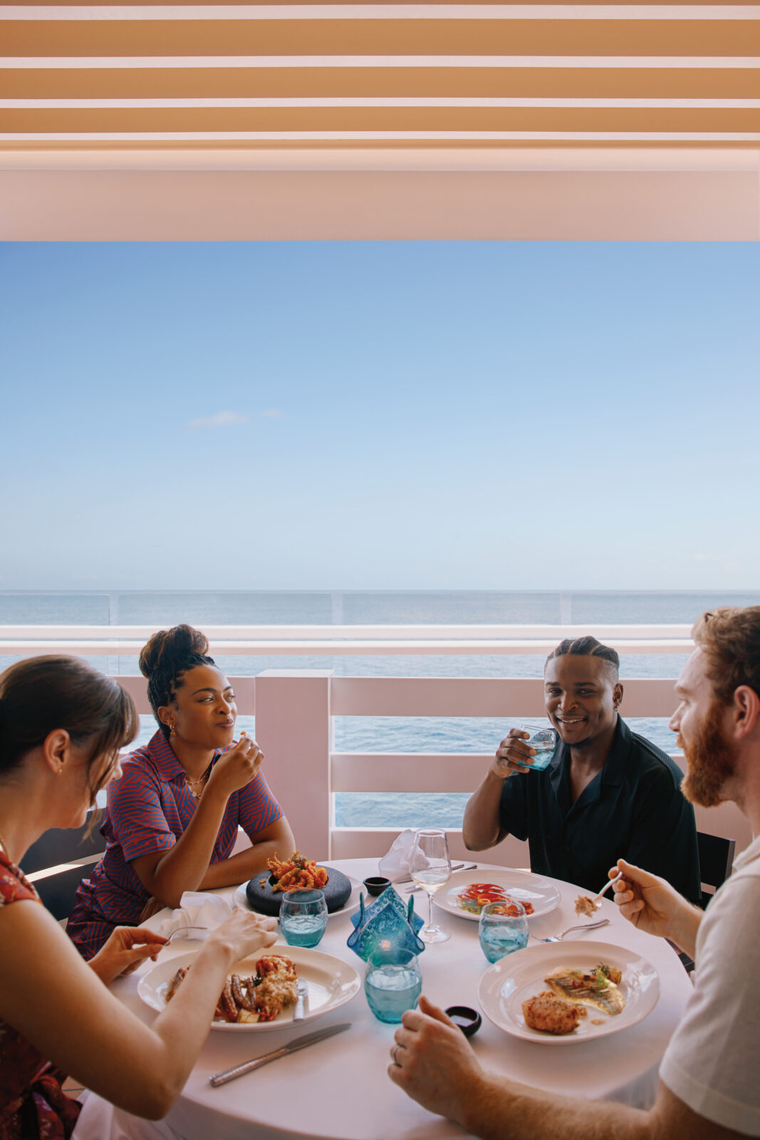 Four people enjoying a meal together on an outdoor cruise ship deck with a clear ocean view.