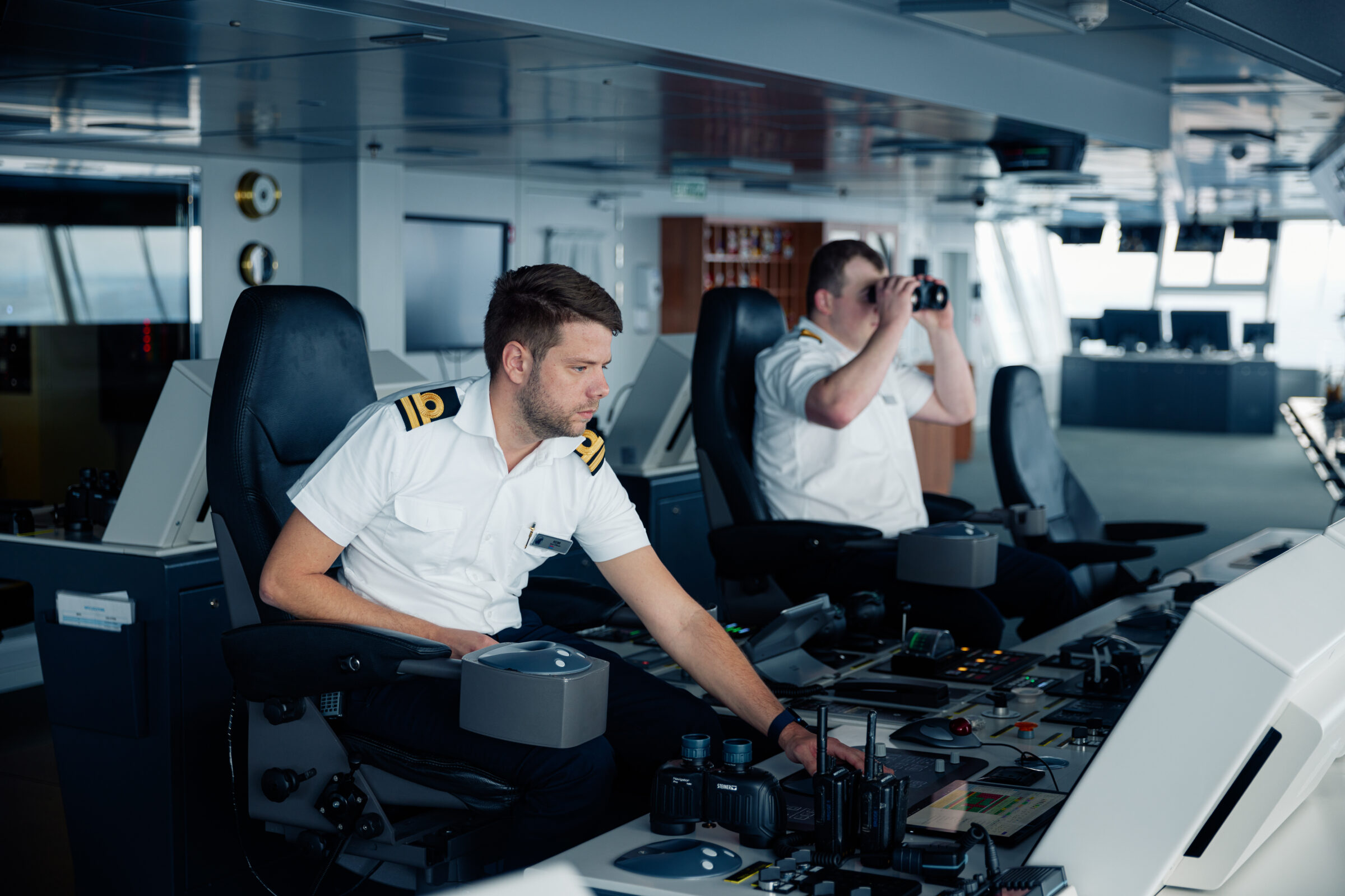 Two ship officers in the bridge of a cruise ship, one using binoculars while the other operates the controls.